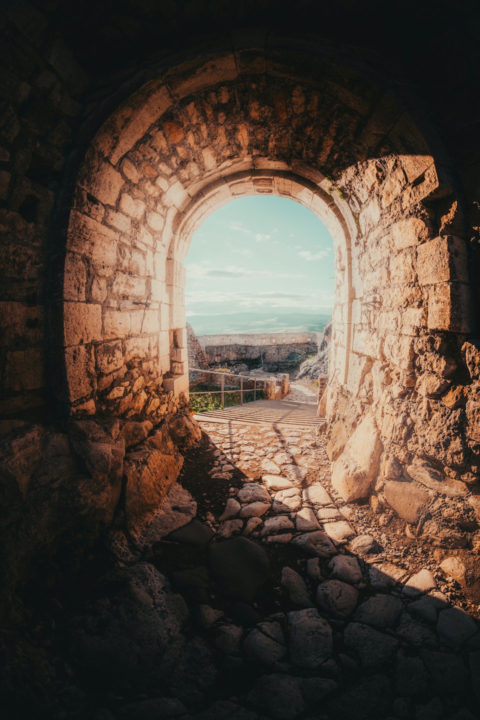 Arco de piedra iluminado por el sol enmarca un camino de adoquines y colinas distantes.